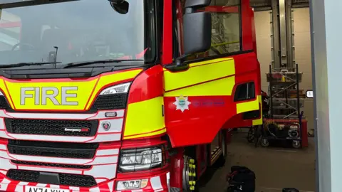 Stock photo of a Cambridgeshire Fire and Rescue engine. It is parked at the Huntingdon station and the doors of the vehicle are open. It is big, red and yellow with the work 'FIRE' on the front in large silver letters. 