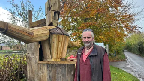 Rob standing beside his Torm Man sculpture standing outside of Torr Cider Farm on an overcast day.