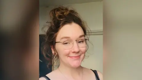 Nadia McVeigh A woman with curly brown and gold hair piled on top of her head. She has gold rimmed glasses, dangly earrings and wears a black vest top.