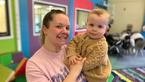 A women with ashy blonde hair in a ponytail. she is wearing a pink t-shirt with blue writing on it. In her arms is a young toddler in a brown knit jumper. They are both looking at the camera smiling.