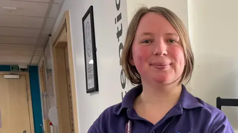 Clare Worden/BBC Elizabeth is a breastfeeding support volunteer. She wears a purple t shirt and NHS lanyard. She has blonde hair tied back and is standing in front of the green reception desk. 