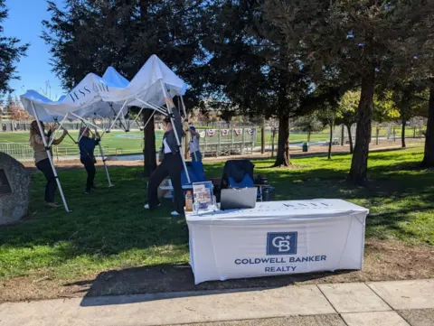 People take down an umbrella behind a table offering realtor services, overlooking a park