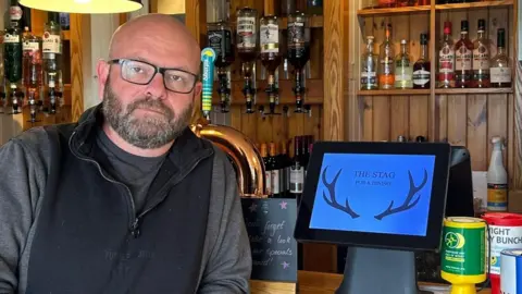 A man with black glasses and a beard leans on a wooden bar with the optics and beer taps behind