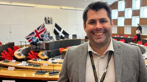 Leigh Frost pictured in the chamber. He is smiling broadly at the camera. He has black hair and is wearing a Cornwall Council lanyard over his striped shirt and grey jacket. Behind him are a couple of councillors getting their papers together. Near Frost's chair is a large England flag and a large St Piran's flag.