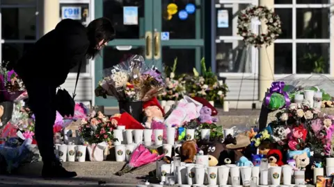 Reuters A woman visits a growing makeshift memorial on the steps of the town hall in Tumbler Ridge. The memorial has a mix of flowers, teddy bears and white cups with small stickers that read "Tumbler Ridge Strong". The woman is wearing black and is leaning forward to read a message left at the memorial.