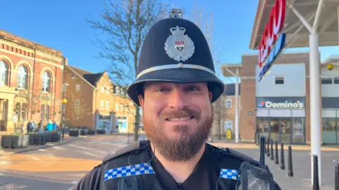 Ash pictured looking into the camera and smiling. He's wearing a black tshirt with a black tactical vest over the top, and a black police helmet with the West Mercia Police badge on it. He's got a police radio attached to his left shoulder. He's stood outside a big Tesco in Madeley, and there's a Dominos pizza shop visible in the distance. The sky is bright blue and clear, with lots of sunshine. 