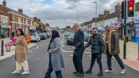 Cllr Shabina in a beige coat and white trousers, Cllr Norin in a grey coat, black headscarf and blue rousers, Cllr Ellis is a grey suit, Cllr Asim in a black puffer jacket and black jeans and Cllr Ali in a brown coat, attempting to cross the road, while looking into the camera.