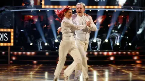 Chris McCausland dan Dianne Buswell dalam setelan jas putih serasi, di tengah tarian di final Strictly