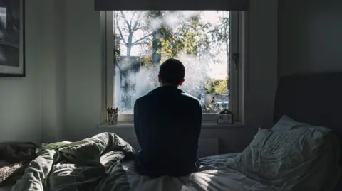 A man looks out of a window. He is in a dark room on an unmade bed. The atmosphere seems negative. 