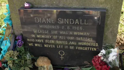 A black grave stone, surrounded by flowers, in the name of Diane Sindall. It has been etched with gold lettering which states: "Diane Sindall murdered 2.8.1986 because she was a woman. In memory of all our sisters who have been raped and murdered. We will never be forgotten."