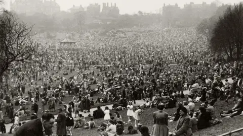 The Harris/Preston City Council A black-and-white photograph, taken from the top of a hill, shows large crowds of people. They are at an outdoor event, with the silhouette of buildings along the skyline in the background.