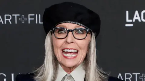Reuters Diane Keaton wearing black-framed sunglasses, a white shirt and a black hate.