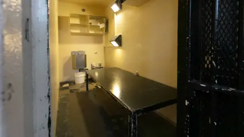 CDCR A photo of one of the cells, with a black table, a few lights, shelves, and a toilet