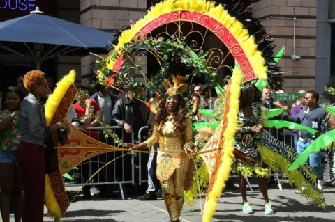 A woman in an elaborate studded yellow costume holds two enormous feather fans with an equally elaborate gold, red and yellow headdress.