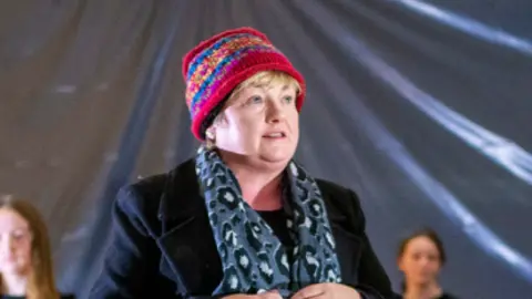 A school play is taking place up at the front is a woman with a hat, black coat and blue patterned scarf. In the background is children wearing a navy top and trousers, or a dress while sitting on a stage.