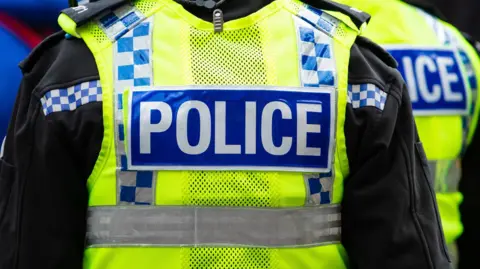 The back of a police officer in uniform with a high vis jacket over the top. Their head is not visible