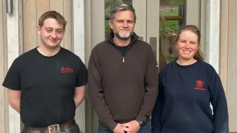 Fiona Callow/BBC Three people stand in front of a modern stonemasonry workshop, two wearing sweaters with the York Minster logo on the chest.