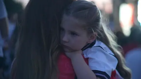 Jamie Niblock/BBC A young blonde girl wearing a white England top clings on to a woman who is wearing a red England top.