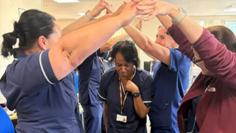 Savitri Cameron A group of people stand in two lines facing each other joining hands together above them to form a 'tunnel' in which Savitri passes underneath in her purple nursing scrubs looking emotional