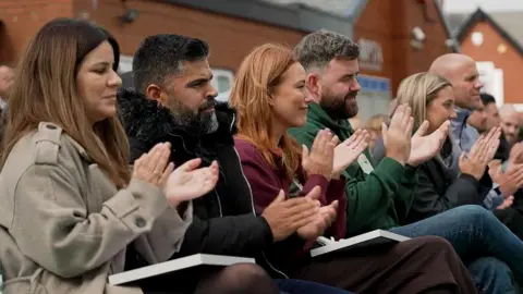 The Southport families are seated outdoors on chairs, clapping with notebooks resting on their laps. Behind them are red brick buildings with pitched roofs.