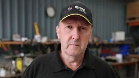 Huw Absalom stands in a workshop wearing a baseball cap and a polo shirt. It is a head and shoulders shot.