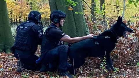 Supplied A photograph of two police officers in woodland. They are wearing protective uniform. One faces away and appears to he holding a gun, the other can be seen from the side, holding a black German Shepherd. They are wearing black clothes.