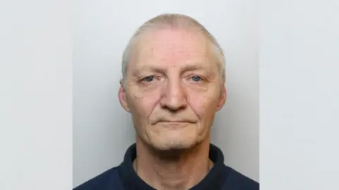 A police custody mugshot of an older man in a blue polo shirt and jumper on a grey background.