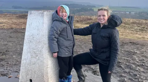Jessica Pryce Arthur, aged 8, wearing a grey coat and a turqoise hoody, smiles at the camera on a hilltop next to his mum, Jessica Pryce, who has wavy blonde hair tied back and is wearing a blue coat. 