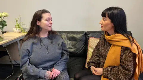 Shariqua Ahmed/BBC Jo, a woman, with long brown hair, wearing a grey jumper, sitting on the sofa next to Theresa, a woman wearing a printed dress and a mustard scarf.