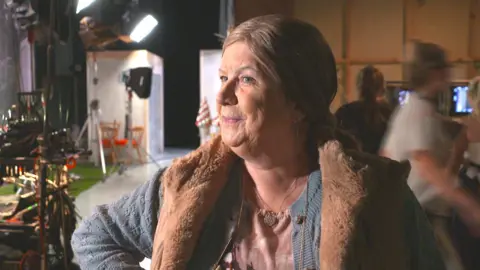 Elaine C Smith, in full character as Christine, gives a cheeky smile on the set of Two Doors Down. We see lights and technical equipment behind her and people watching TV monitors
