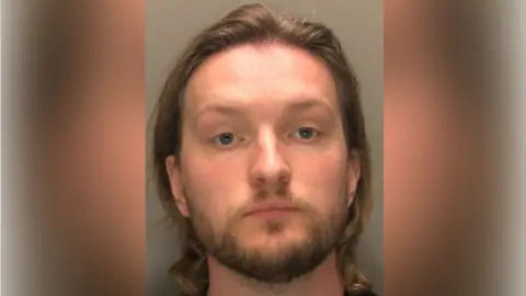 A police custody shot of Rhys Lynn. He has long brown hair and brown facial hair. The wall behind him is grey.