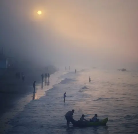 @jonski_the_wanderer People playing in the sea on a misty day with the sun a disc as seen through cloud