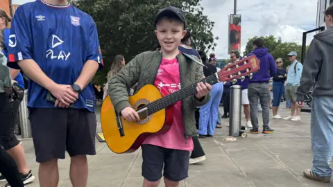 Zoie O'Brien/BBC A boy holds an acoustic guitar. He wears a dark cap, a green coat, a pink Ipswich Town shirt and shorts.