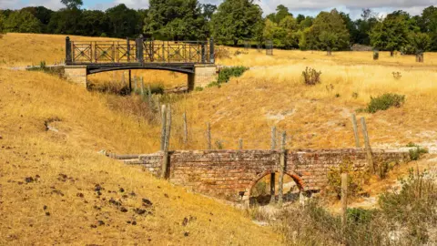 PA Media Two bridges, one in the foreground and one in the distance, both stretch across two fields which have dried grass and hay. Several green trees are in the distance.