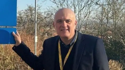 Councillor Ed O'Driscoll wearing a dark jacket, is facing the camera, touching a large sign outside Ross-on-Wye Community Hospital.