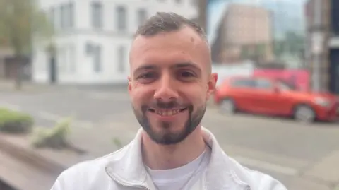 A man with short brown hair and a short brown beard and moustache smiles at the camera. He wears a white quarter zip jumper with a white t-short underneath. 