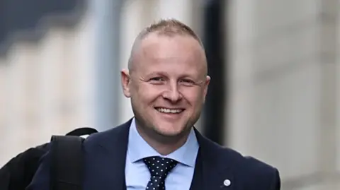 Jamie Bryson is wearing a light blue shirt and a navy and white spotted tie. He is also wearing a navy blazer. He has a black rucksack over one shoulder. The background is blurry. 