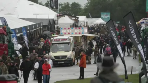 The image shows a busy outdoor event with crowds of people walking between rows of tents, stalls and large vehicles. A food van with a sign reading “Kelly Whip” is parked along the main path, while flags, branded banners and exhibition stands line both sides, suggesting a large agricultural show or fair.