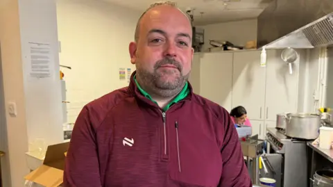 Paul McCusker in a red jumper with a green t-shirt. He is standing in front of a kitchen. He has a black beard and black hair.
