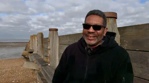 BBC Fred Jackson stands on a beach in front of a large wooden structure and with the sea visible in the background. He is wearing dark sunglasses and a black zip up hoodie.