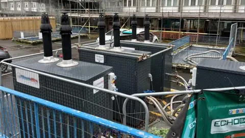 George Carden/BBC Temporary large metal boilers with chimneys on the top in the car park of the flats