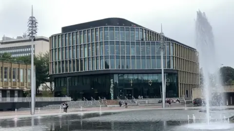 BBC A shiny glass multi storey office building, in a city square with a large fountain in front of it.