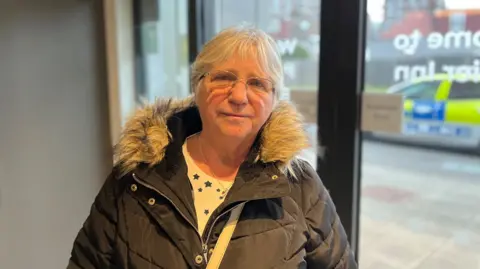 A woman with glasses and a black winter coat stands indoors near a glass door. A police car is visible outside.