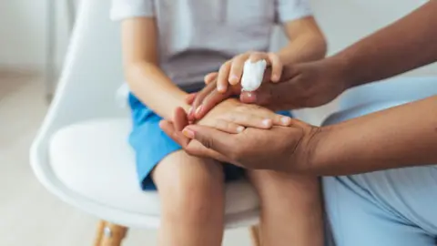 A stock image of a child sitting on a chair, seen only from the neck down and holding the hands of an adult whose arms only we can see. One of the child's fingers has a bandage on it.