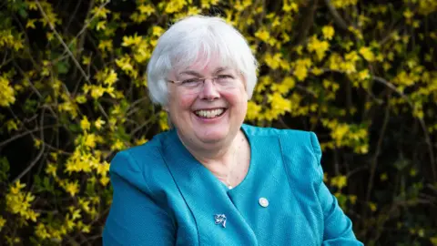 Alice Chapman Photography Charlotte Cane stares at the camera. She is smiling and wears a light blue jacket with two small metal pin badges. In the background is a yellow forsythia bush.