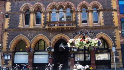 The front view of Hereford Museum and Art Gallery. There are bikes stacked in a bike rack and five large arched windows. There three flower hanging baskets attached to the windows.