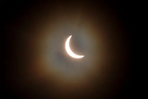 Getty Images The sun is pictured during a partial solar eclipse. The sky is dark and cloudy. The sun is a thin arc in the sky.