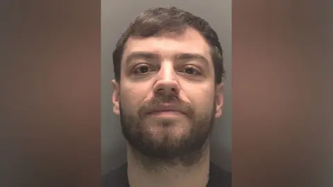 A police mugshot of Lee Harrison, 27, who has brown hair and a brown beard and smirks at the camera.