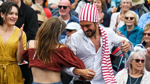 A man wearing a stripey head scarf and a white shirt dances with a woman wearing a red crop top. Their arms are linked. There is a crowd of people behind them.