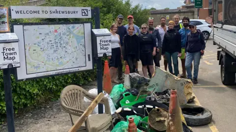 Shrewsbury Vegans A group of people standing beside a sign which reads Welcome to Shrewsbury and a large pile of rubbish, including a chair, two traffic cones, a plank of wood, a tyre and plastic boxes containing other waste